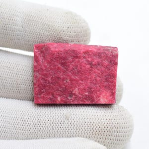 Peut inclure: Un morceau rectangulaire de pierre précieuse rhodonite brute avec une couleur rose magenta vibrante et une surface rugueuse et texturée. La pierre présente des variations naturelles de teinte et des inclusions minérales.