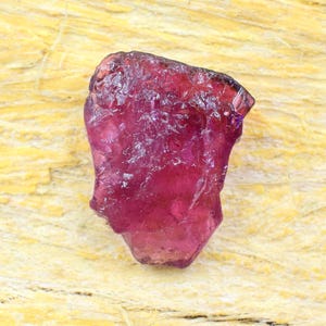 Puede incluir: Una piedra preciosa en bruto y sin pulir con un tono magenta profundo y rojo translúcido. La piedra presenta una superficie irregular y facetada con crestas y hendiduras naturales, mostrando un color vibrante y saturado en su estructura.
