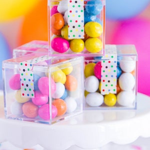 May include: Three clear square boxes filled with colorful candy. The boxes are stacked on a white cake stand. The boxes are decorated with colorful polka dot washi tape.