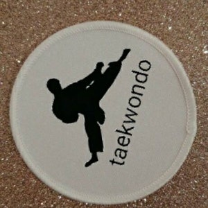 Puede incluir: Parche redondo blanco con una silueta negra de una persona realizando una patada de taekwondo. El texto "taekwondo" está escrito verticalmente en el lado derecho del parche.