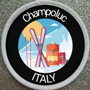 May include: Round patch with the text "Champoluc ITALY". The patch features a ski scene with skis, poles, a backpack, and ski boots. The background includes a sun, mountains, and a blue sky.