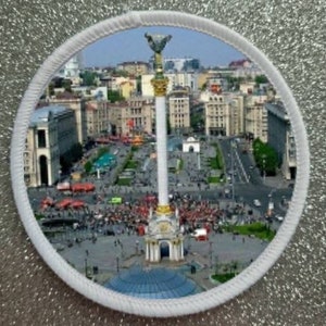 May include: A round patch featuring a detailed image of a city square with a tall white column topped with a gold statue. The patch is bordered by a white, textured trim and set against a glittery silver background.