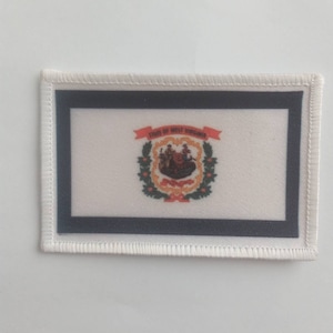 Könnte beinhalten: Weißer gestickter Aufnäher mit schwarzem Rand, der das Staatssiegel von West Virginia zeigt. Das Siegel zeigt einen Bauern, der eine Garbe Weizen hält, einen Bergmann, der eine Spitzhacke hält, und einen Holzfäller, der eine Axt hält. Das Siegel ist von einem Lorbeerkranz und einem roten Band mit der Aufschrift "State of West Virginia" in Weiß umgeben.