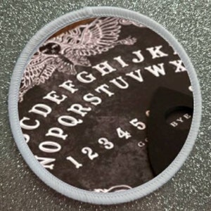 Op de afbeelding: Een ronde zwarte en witte patch met een witte rand. De patch heeft een vintage-stijl Ouija-bord met de letters CDEFGHIJKLMNOPQRSTUVWXYZ, de nummers 12345 en het woord "BYE" op de planchette.