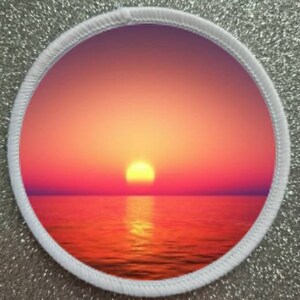 May include: A round patch with a white border and a sunset over the ocean. The sky is a gradient of pink, orange, and yellow. The sun is setting in the distance, casting a golden glow on the water.
