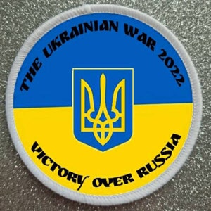 May include: A white circular patch with a blue and yellow Ukrainian flag in the center. The flag features the Ukrainian coat of arms, a trident. The text "THE UKRAINIAN WAR 2022 VICTORY OVER RUSSIA" is written around the flag.