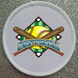 Peut inclure: Écusson rond blanc avec un ballon de softball, deux batte de baseball croisées et le mot "SOFTBALL" en lettres bleues et rouges sur un losange vert et blanc.