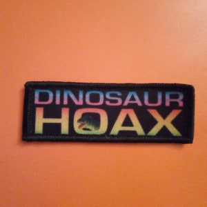 May include: Black embroidered patch with the text "Dinosaur Hoax" in rainbow colours.