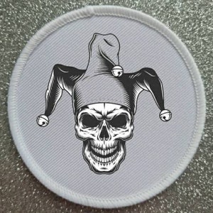May include: A white round patch featuring a black and white skull wearing a jester's hat with bells.