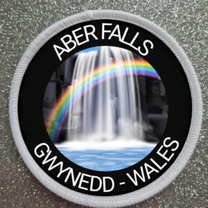 May include: A white round patch with black text and a picture of a waterfall with a rainbow. The text reads "Aber Falls Gwynedd - Wales".
