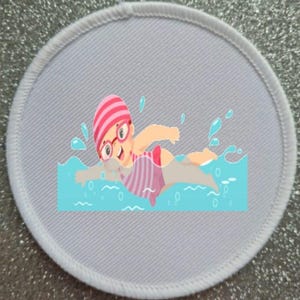 May include: A white oval patch with a cartoon illustration of a person swimming in blue water. The person is wearing a pink and white striped swimsuit and a pink swim cap. The person is wearing glasses and has a big smile.