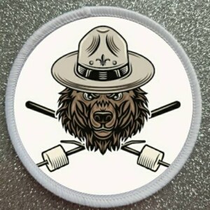 May include: A white round patch featuring a brown bear wearing a tan ranger hat with a black band and a fleur-de-lis design. The bear has a serious expression and is holding two crossed sticks with marshmallows on the ends.