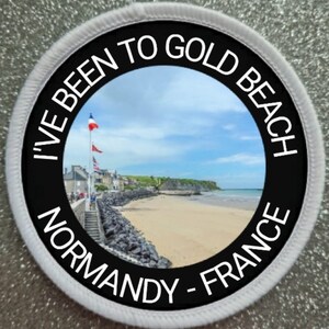Könnte beinhalten: Ein runder weißer und schwarzer Aufnäher mit dem Text "I've Been To Gold Beach Normandy - France" um ein Foto eines Strandes mit einer Stadt im Hintergrund.