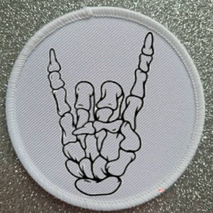 May include: White round patch with a black line drawing of a skeleton hand making the rock and roll hand gesture.