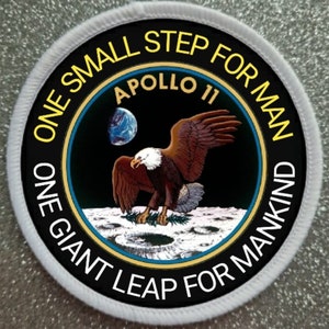 Pode incluir: Um remendo circular branco e preto com o texto "One small step for man, one giant leap for mankind" ao redor da borda. O centro do remendo apresenta uma águia careca pousando na lua com o texto "Apollo 11" acima.