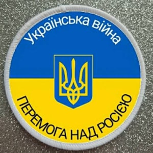 Puede incluir: Un parche circular blanco con colores azul y amarillo, que representa la bandera ucraniana con el escudo de armas del país, un tridente, en el centro. El texto "Українська війна Перемога над Росією" está escrito alrededor de la bandera en negro.