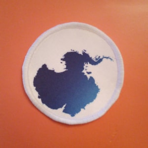 May include: A white round patch with a blue and teal gradient silhouette of Antarctica.
