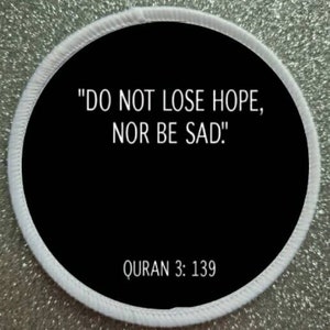 Puede incluir: Un parche redondo blanco y negro con el texto "DO NOT LOSE HOPE, NOR BE SAD!" y "QURAN 3: 139" en blanco.