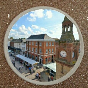 May include: A circular patch featuring a town square scene with buildings and a clock tower. The image is framed by a white border and set against a glittery, gold-toned background. People are walking in the square under a blue sky.