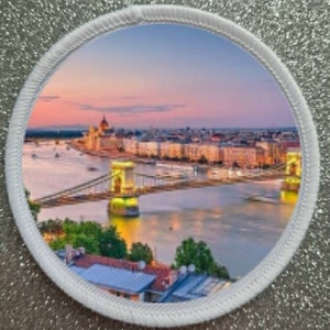 May include: A white circular patch featuring a colourful image of the Chain Bridge in Budapest, Hungary. The bridge is illuminated at night and the city skyline can be seen in the background. The sky is a vibrant pink and orange.