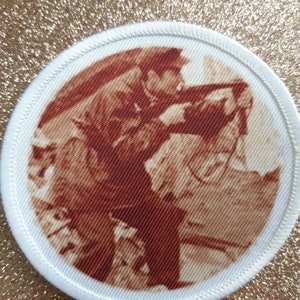 May include: A white circular patch with a brown image of two people in a vintage photograph. The image is of a man and a woman, with the man holding the woman's hand. The image is slightly blurry.