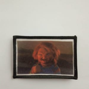 Könnte beinhalten: Ein gestickter schwarzer und weißer Aufnäher mit einer Nahaufnahme des Gesichts einer Puppe mit einem bedrohlichen Ausdruck. Die Puppe trägt ein blau-weiß gestreiftes Hemd.