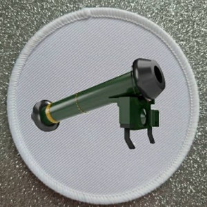 May include: A white round patch with a green and black toy rocket launcher. The rocket launcher has a black barrel and a green body with a gold band around the base.