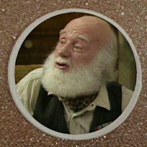 Può includere: Primo piano di un uomo con barba e baffi bianchi, che indossa un gilet marrone e una sciarpa a motivi. Sta guardando a sinistra e ha un'espressione sorpresa sul viso.