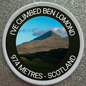 May include: White round patch with black text and a photo of a mountain. The text reads "I've Climbed Ben Lomond 974 Metres - Scotland".