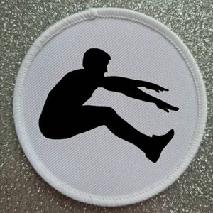 Peut inclure: Écusson rond avec une silhouette noire d'un athlète en plein saut en longueur. Le patch a un fond blanc et une bordure blanche cousue. L'image est placée sur un fond argenté scintillant.
