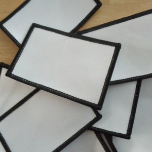 May include: A collection of rectangular white patches with black stitched borders. The patches are blank, offering a clean canvas for customization. The black stitching provides a defined edge to the white fabric.