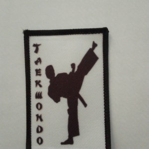 Può includere: Una toppa bianca con bordo nero, che presenta una silhouette marrone di una persona che esegue un calcio di arti marziali. La parola "TAEKWONDO" è scritta verticalmente in marrone sul lato sinistro della toppa.