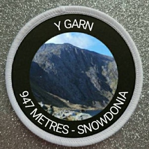 May include: A black and white round patch with a white border. The patch features a photo of a mountain range with the text "Y Garn 947 Metres - Snowdonia" in white.