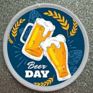 Könnte beinhalten: Ein runder weißer Patch mit blauem Hintergrund und einem Design aus zwei Bierkrügen mit Schaum oben drauf. Die Krüge sind mit gelbem Bier gefüllt und stoßen an. Der Text "Beer Day" ist in weißen Buchstaben unter den Krügen geschrieben.