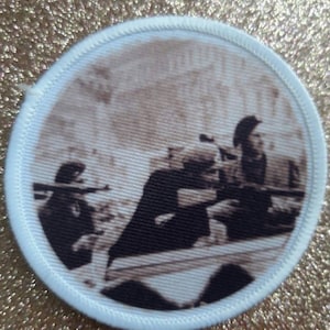 May include: A white circular patch with a black and white image of three people in a military setting. The image is faded and has a vintage look.