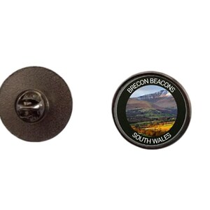 Op de afbeelding: Een ronde metalen speld met een fotografisch landschap van de Brecon Beacons in Zuid-Wales. De tekst 'BRECON BEACONS' en 'SOUTH WALES' is in het wit gedrukt op een zwarte rand. De achterkant toont een standaard metalen vlindersluiting.