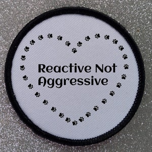 May include: White round patch with black border. The patch has a heart shape made of paw prints and the text "Reactive Not Aggressive" inside the heart.