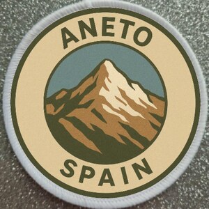 Puede incluir: Parche circular con la inscripción "ANETO SPAIN" y una ilustración de montaña. El diseño presenta un pico montañoso en marrón y blanco, sobre un cielo azul, dentro de un borde beige y un borde cosido blanco.