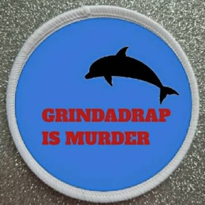 May include: A white circular patch with a blue background and a black silhouette of a dolphin jumping out of the water. The text "GRINDADRAP IS MURDER" is written in red letters below the dolphin.