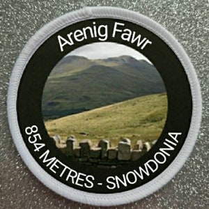 May include: Round patch with a black border and white stitching. The patch features a landscape image of a mountain range with the text "Arenig Fawr" at the top and "854 METRES - SNOWDONIA" at the bottom.