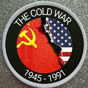 Può includere: Un cerotto circolare bianco con un cerchio nero al centro. Il cerchio nero ha il testo "THE COLD WAR" in lettere bianche attorno al bordo. All'interno del cerchio nero c'è un simbolo comunista rosso e giallo e una bandiera americana strappata. Il testo "1945 - 1991" è stampato in bianco nella parte inferiore del cerotto.