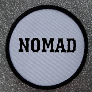 Op de afbeelding: Ronde witte patch met zwarte rand en het woord "NOMAD" in zwarte letters.