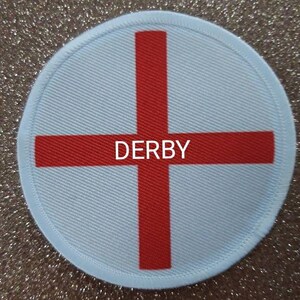 Könnte beinhalten: Runder weißer Patch mit einem roten Kreuz und dem Wort "DERBY" in der Mitte.