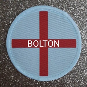 Puede incluir: Un parche redondo blanco y rojo con una cruz y la palabra "BOLTON" en el centro.