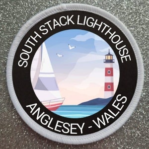 May include: A white round patch with black text and a black circle in the centre. The text reads "South Stack Lighthouse Anglesey - Wales". The circle contains a colourful illustration of a lighthouse, a sailboat, and seagulls against a blue sky.