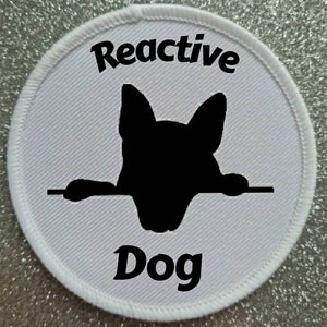 Peut inclure: Patch rond blanc avec du texte noir et une silhouette de chien qui regarde par-dessus une clôture. Le texte indique "Reactive Dog".