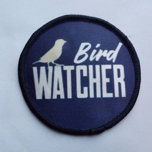 Puede incluir: Un parche redondo azul y blanco con una silueta de pájaro blanca y el texto "Bird Watcher" en blanco.