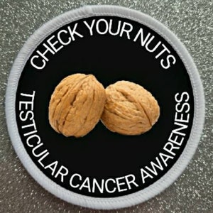 Puede incluir: Parche redondo blanco con texto negro que dice "Check Your Nuts Testicular Cancer Awareness". Dos nueces están en el centro del parche.