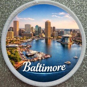Peut inclure: Un écusson rond représentant le port intérieur de Baltimore, avec la ligne d'horizon de la ville, des bateaux et de l'eau bleue. Le mot "Baltimore" est écrit en blanc. L'écusson a une bordure blanche.