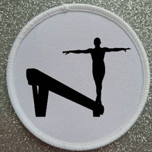 Peut inclure: Un écusson rond blanc avec une silhouette noire d'un gymnaste effectuant un exercice sur une poutre d'équilibre.